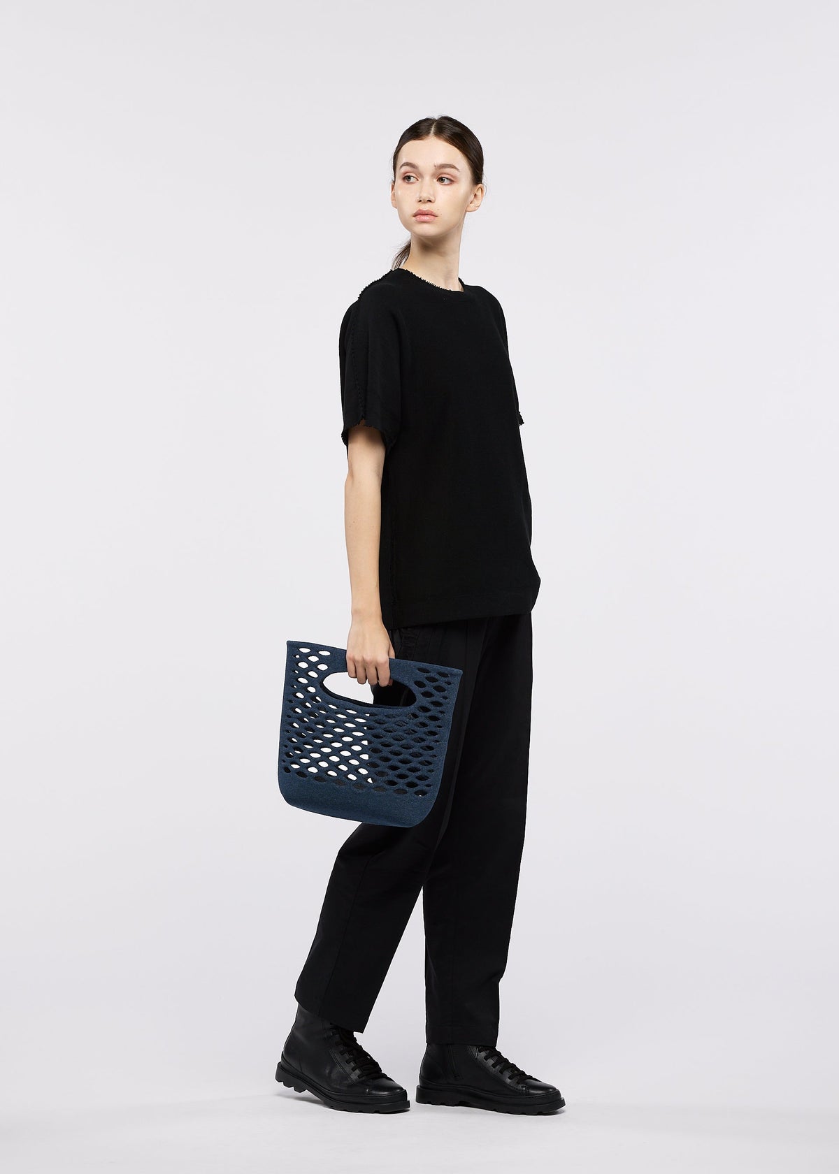 A person in loose black clothing stands against a white background, holding the navy blue MOKKO WASHI tote bag by GOOD GOODS ISSEY MIYAKE and looking to the side. view 7 of 8