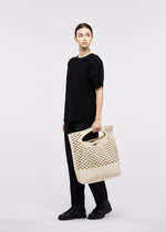 A person stands against a plain background, wearing a loose black outfit and shoes, holding the large beige MOKKO WASHI tote bag with cut-out handles from GOOD GOODS ISSEY MIYAKE.