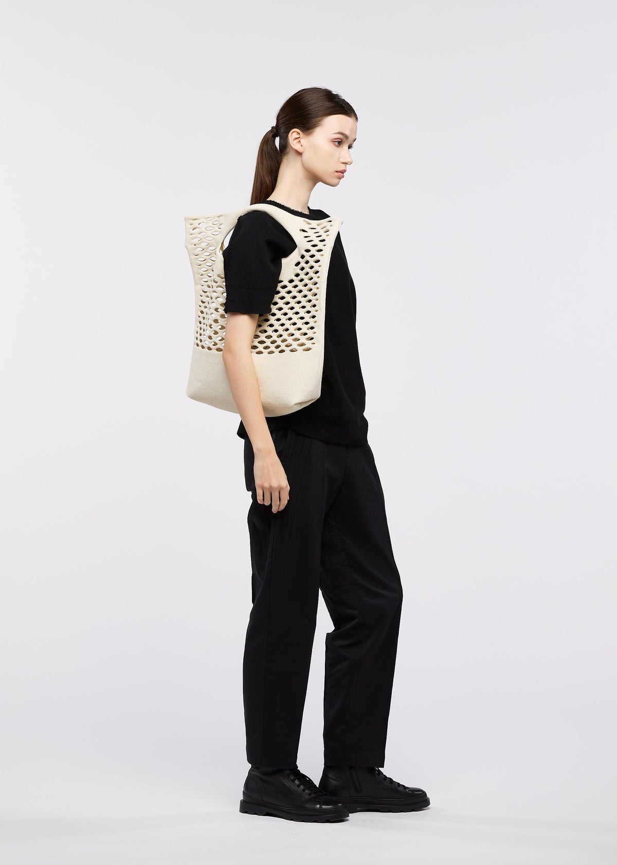 A woman with straight brown hair, in black pants and a black top, stands sideways against a plain light background, carrying the cream MOKKO WASHI tote bag by GOOD GOODS ISSEY MIYAKE over her shoulder—a perfect summer accessory. view 9 of 10