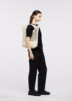 A woman with straight brown hair, in black pants and a black top, stands sideways against a plain light background, carrying the cream MOKKO WASHI tote bag by GOOD GOODS ISSEY MIYAKE over her shoulder—a perfect summer accessory.