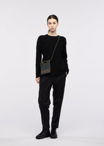 A person stands against a plain background wearing a black sweater, black pants, black shoes, and the MOKKO WALE crossbody bag from GOOD GOODS ISSEY MIYAKE, with a neutral expression and relaxed posture.