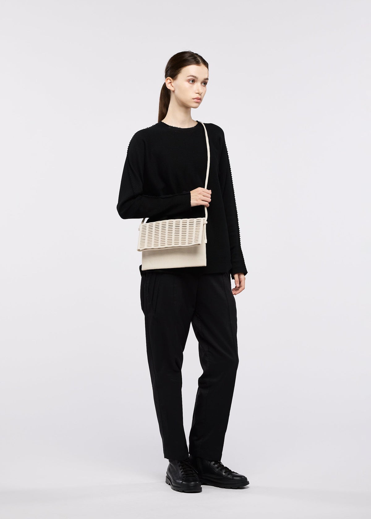 A person stands against a white background in all-black attire, holding the beige textured shoulder bag MOKKO WALE by GOOD GOODS ISSEY MIYAKE. view 11 of 13