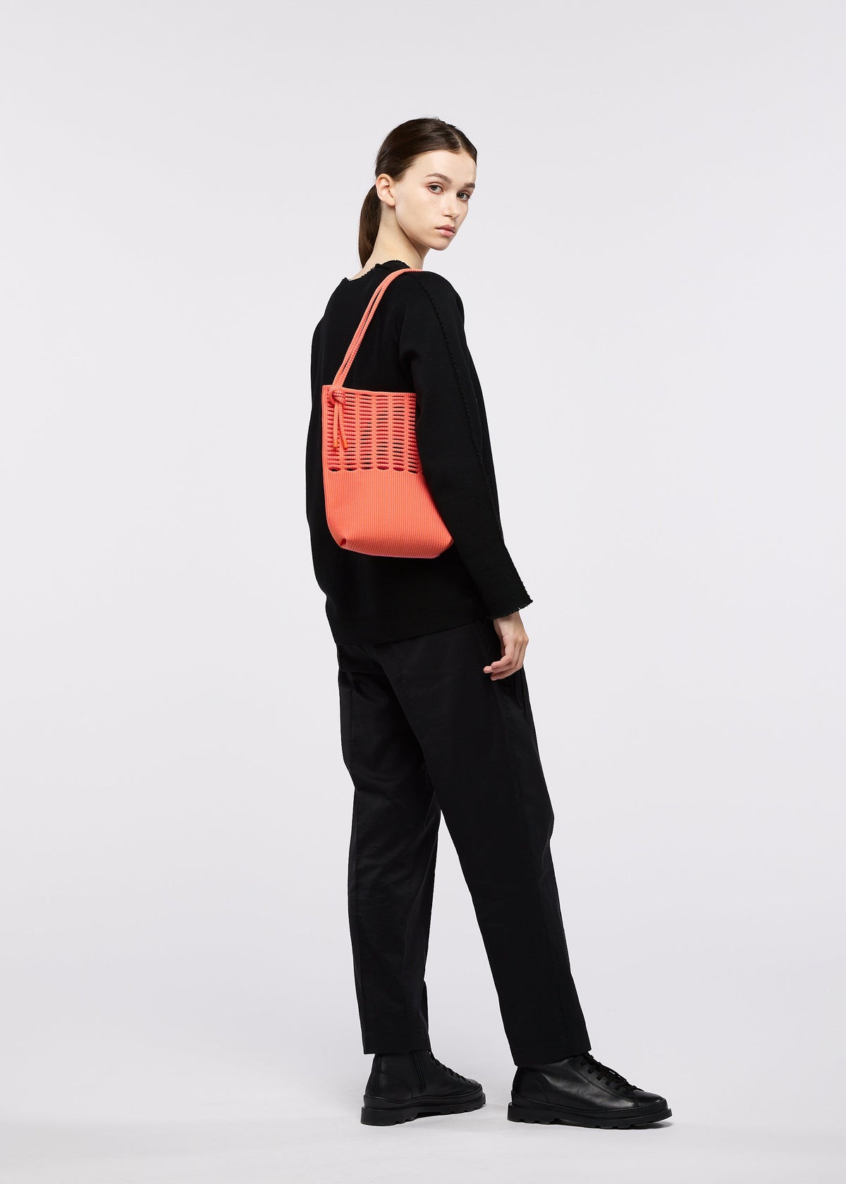A woman in black clothes carries the MOKKO WALE by GOOD GOODS ISSEY MIYAKE, a coral-striped shoulder bag, as she looks over her shoulder against a plain white background. view 12 of 13