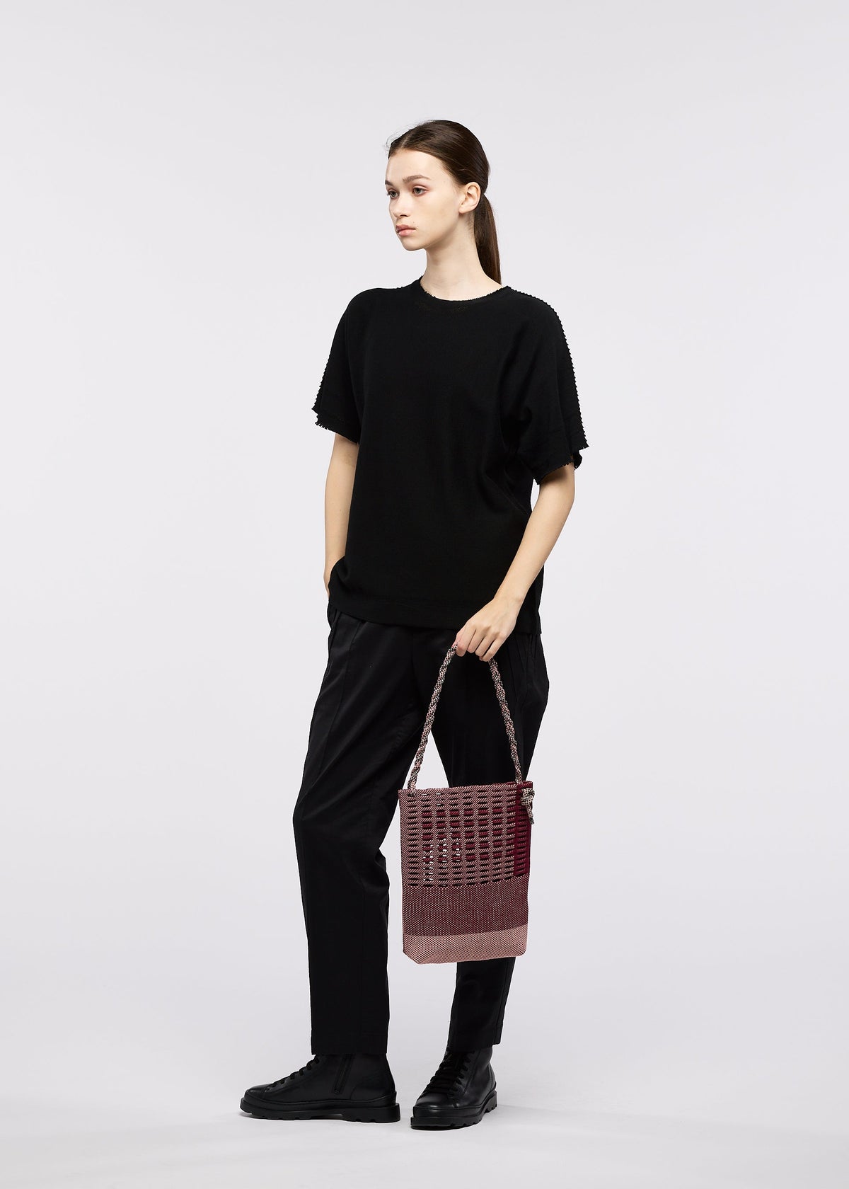 A woman stands against a plain white background, dressed in black and holding the GOOD GOODS ISSEY MIYAKE MOKKO KNOT shoulder bag with both hands while gazing slightly to the side. view 8 of 10