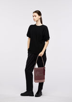 A woman stands against a plain white background, dressed in black and holding the GOOD GOODS ISSEY MIYAKE MOKKO KNOT shoulder bag with both hands while gazing slightly to the side.