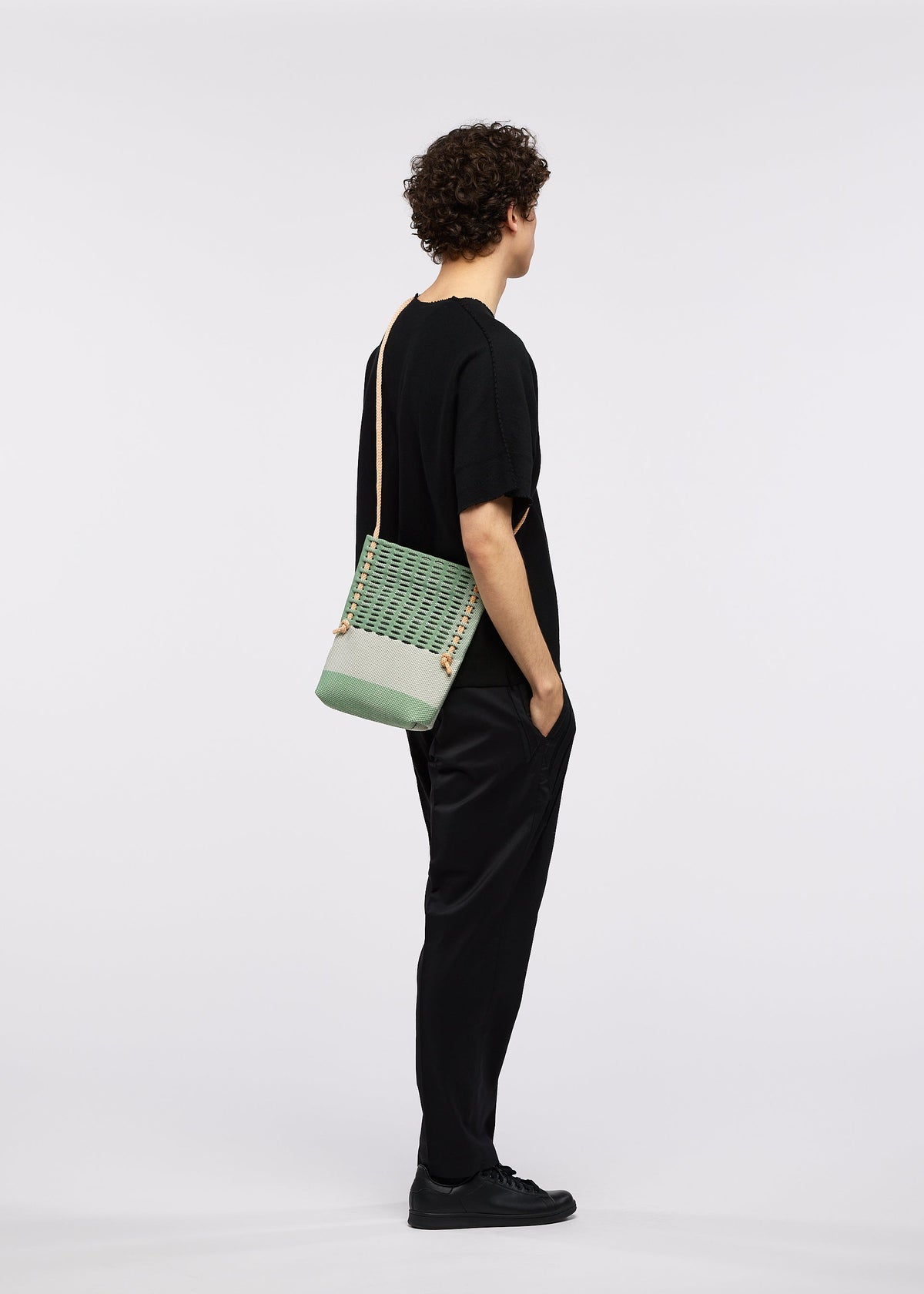 A person with curly hair, dressed in black with black sneakers, stands in profile with hands in pockets, wearing the GOOD GOODS ISSEY MIYAKE MOKKO KNOT adjustable cord bag against a plain white background. view 9 of 10