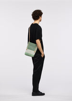 A person with curly hair, dressed in black with black sneakers, stands in profile with hands in pockets, wearing the GOOD GOODS ISSEY MIYAKE MOKKO KNOT adjustable cord bag against a plain white background.