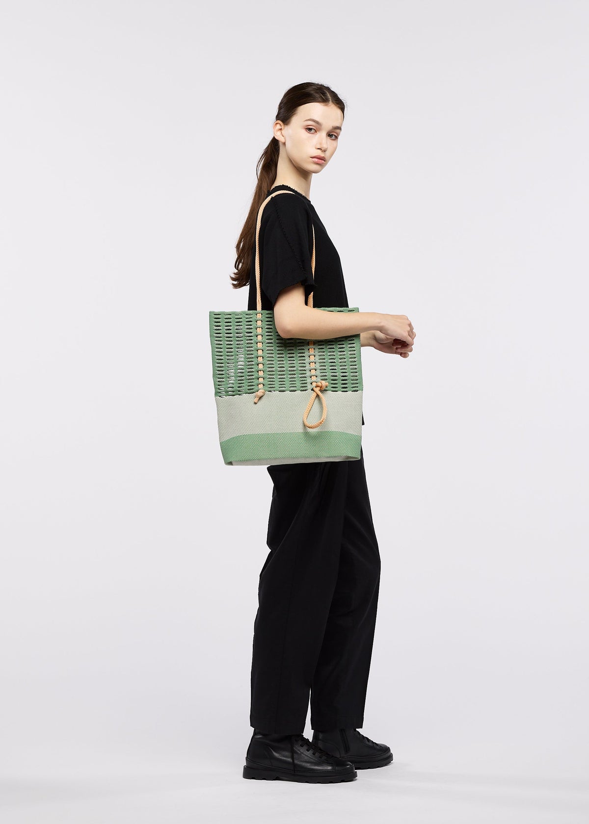 A young woman with long brown hair in a ponytail stands sideways in black attire and shoes, holding the green and beige MOKKO KNOT bag with rope handles by GOOD GOODS ISSEY MIYAKE against a plain white background. view 8 of 10