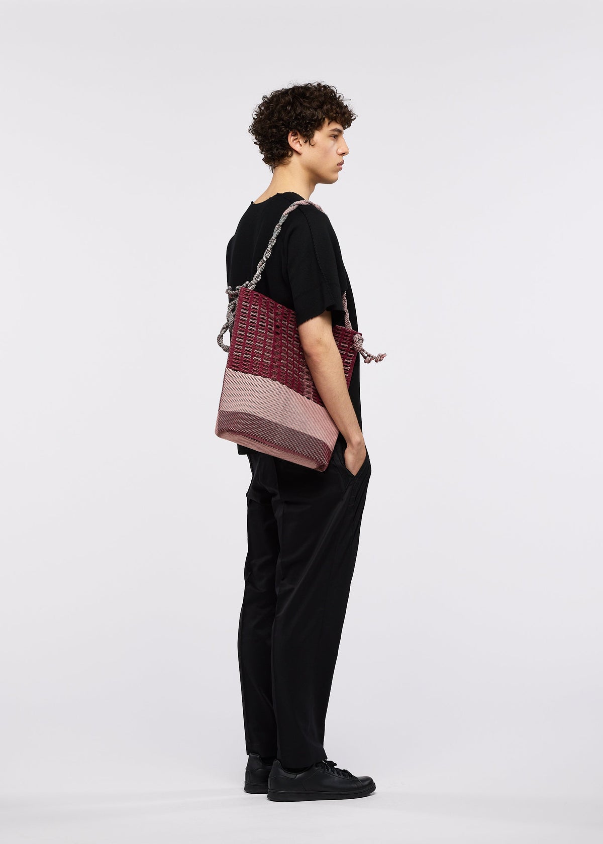A person with curly hair, in black clothes and shoes, stands in profile against a white background, carrying the large maroon and pink MOKKO KNOT bag by GOOD GOODS ISSEY MIYAKE over one shoulder. view 9 of 10