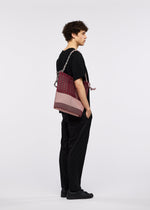 A person with curly hair, in black clothes and shoes, stands in profile against a white background, carrying the large maroon and pink MOKKO KNOT bag by GOOD GOODS ISSEY MIYAKE over one shoulder.