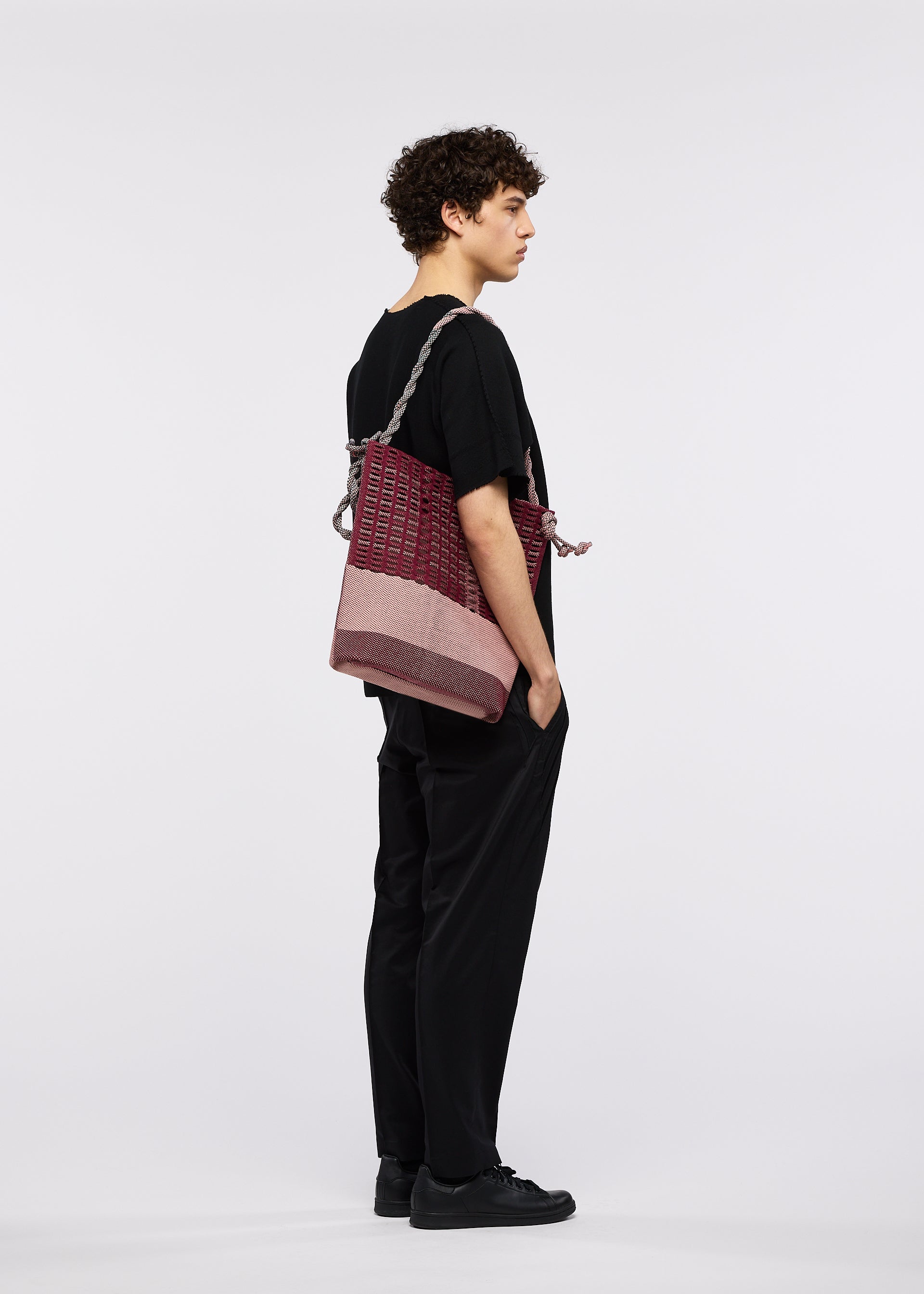 Zoomed view A person with curly hair, in black clothes and shoes, stands in profile against a white background, carrying the large maroon and pink MOKKO KNOT bag by GOOD GOODS ISSEY MIYAKE over one shoulder.