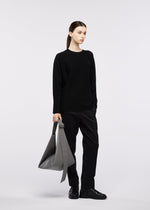A woman with brown hair in a ponytail stands against a light gray background, holding the GOOD GOODS ISSEY MIYAKE LEAF large gray suede-like shoulder bag. She wears a black sweater, pants, and shoes.