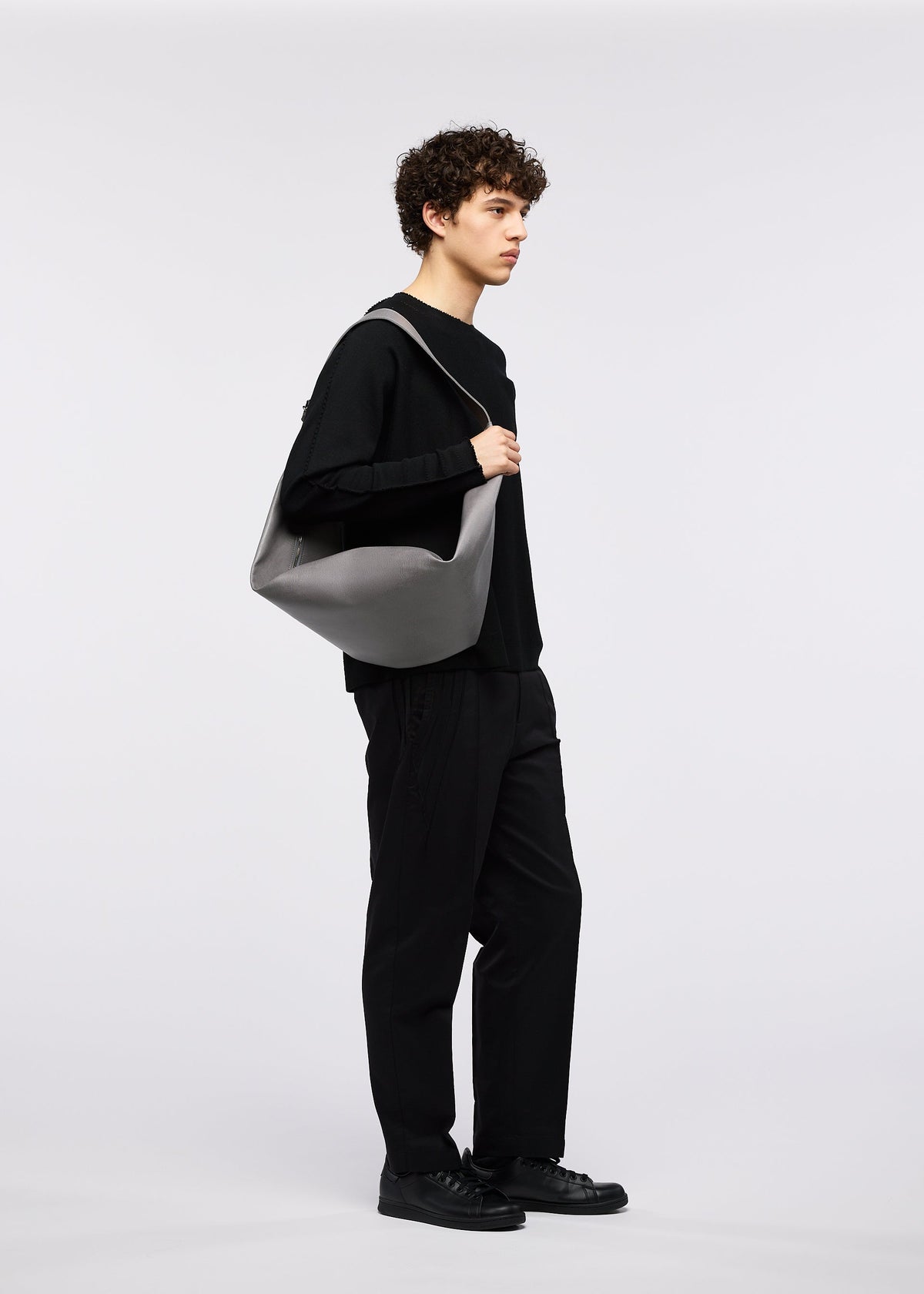 A person with curly hair in a black outfit and shoes stands sideways, holding the gray LEAF shoulder bag by GOOD GOODS ISSEY MIYAKE against a plain white background. view 9 of 9