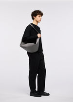 A person with curly hair in a black outfit and shoes stands sideways, holding the gray LEAF shoulder bag by GOOD GOODS ISSEY MIYAKE against a plain white background.