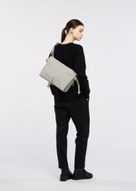 A person in black clothing and boots stands facing away, wearing the GOOD GOODS ISSEY MIYAKE JABARA light gray shoulder bag featuring an accordion pleat design over their shoulder against a plain white background.