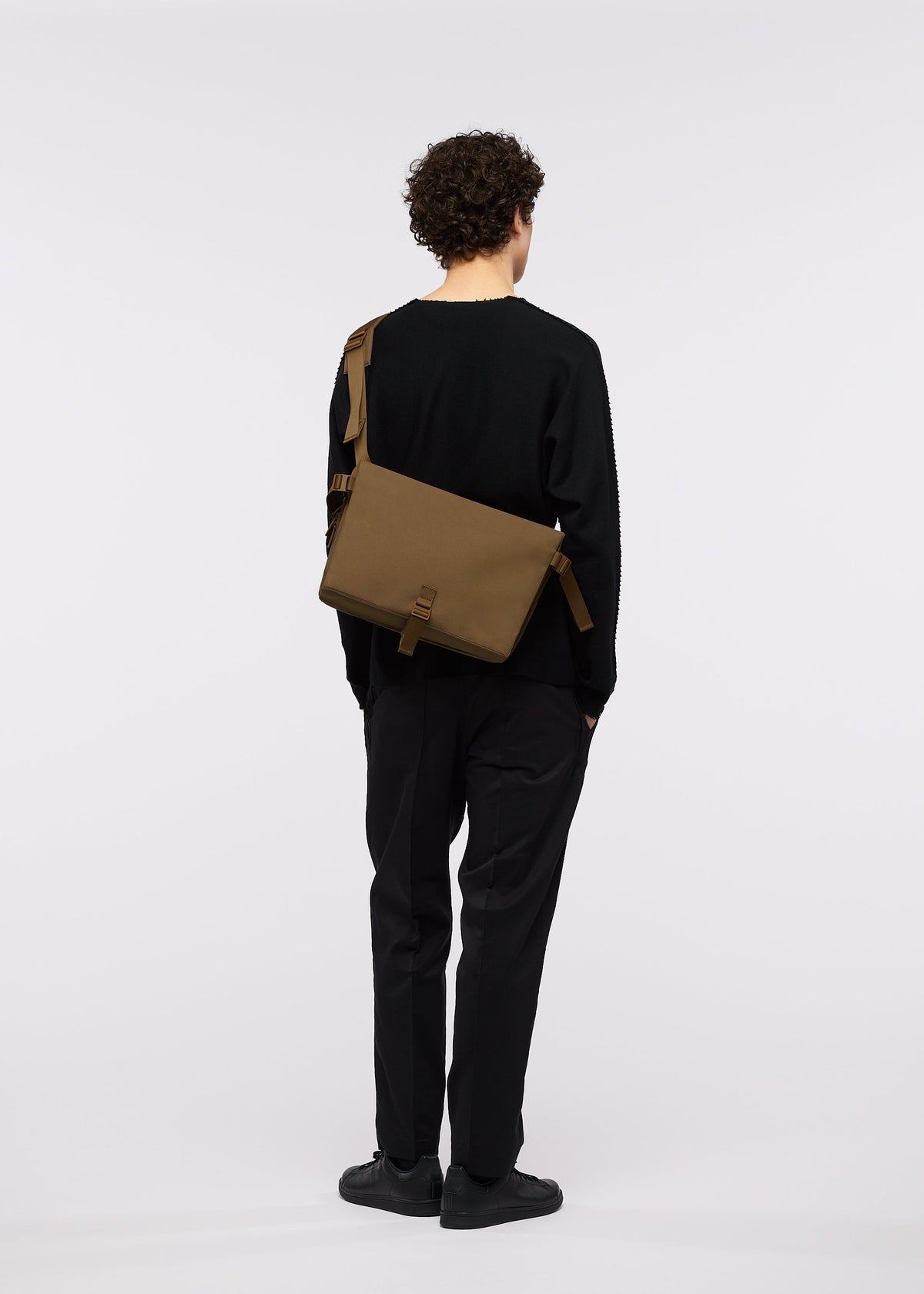 A person with curly hair, dressed in black, stands facing away against a plain white background. They wear the GOOD GOODS ISSEY MIYAKE JABARA brown accordion pleat bag slung crossbody over their shoulder. view 11 of 11