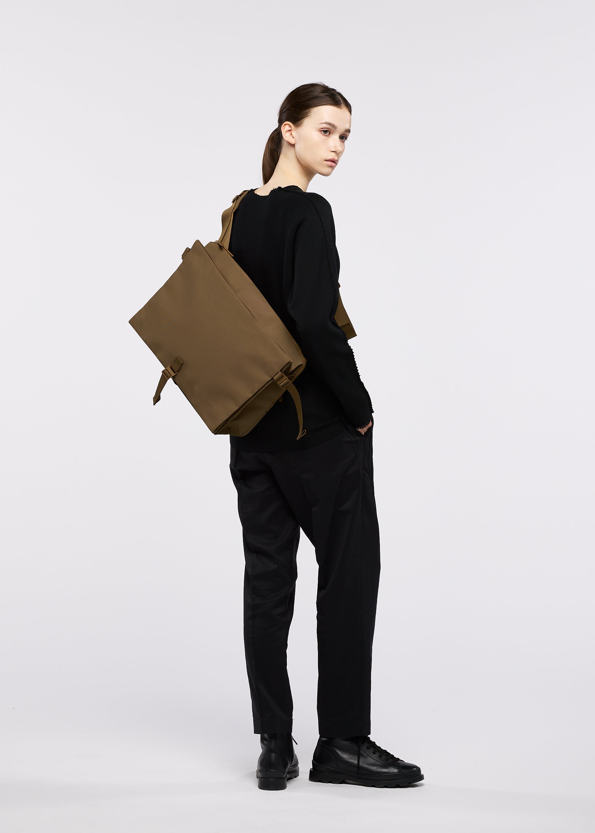 Zoomed view A person with dark hair pulled back stands against a white background, dressed in a black sweater, black trousers, and black boots. Slung over their shoulder is the large, structured JABARA shoulder bag by GOOD GOODS ISSEY MIYAKE in brown, featuring a convenient laptop pocket.