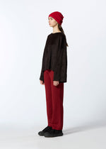 A person stands sideways against a plain light background, wearing HaaT’s KUMO SHIBORI KNIT red cap, a loose dark brown sweater, red wide-leg recycled polyester pants, and black shoes.