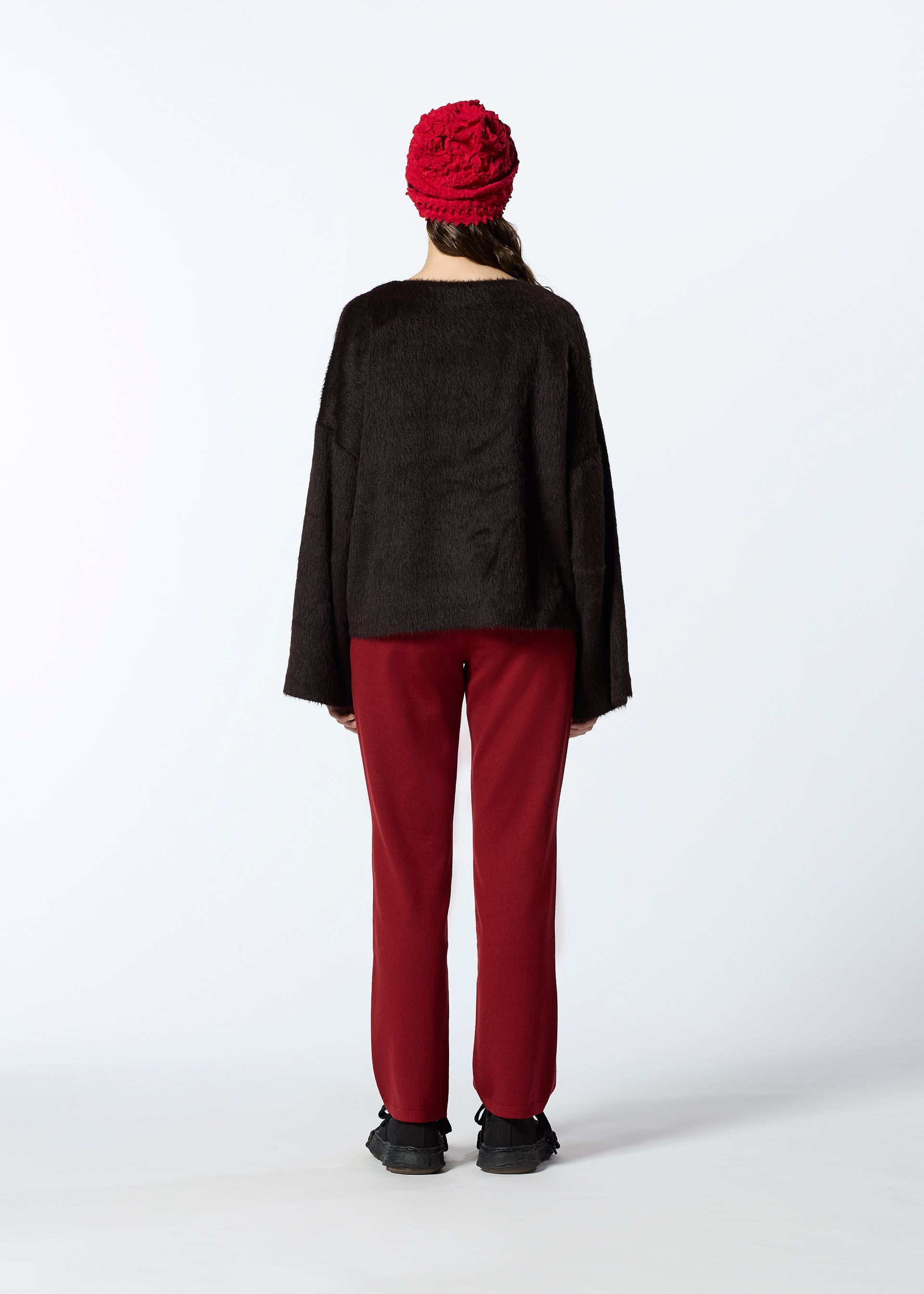 Zoomed view A person faces away, wearing a dark brown long-sleeve top, red pants, black shoes, and the HaaT KUMO SHIBORI KNIT cap. The background is plain white.