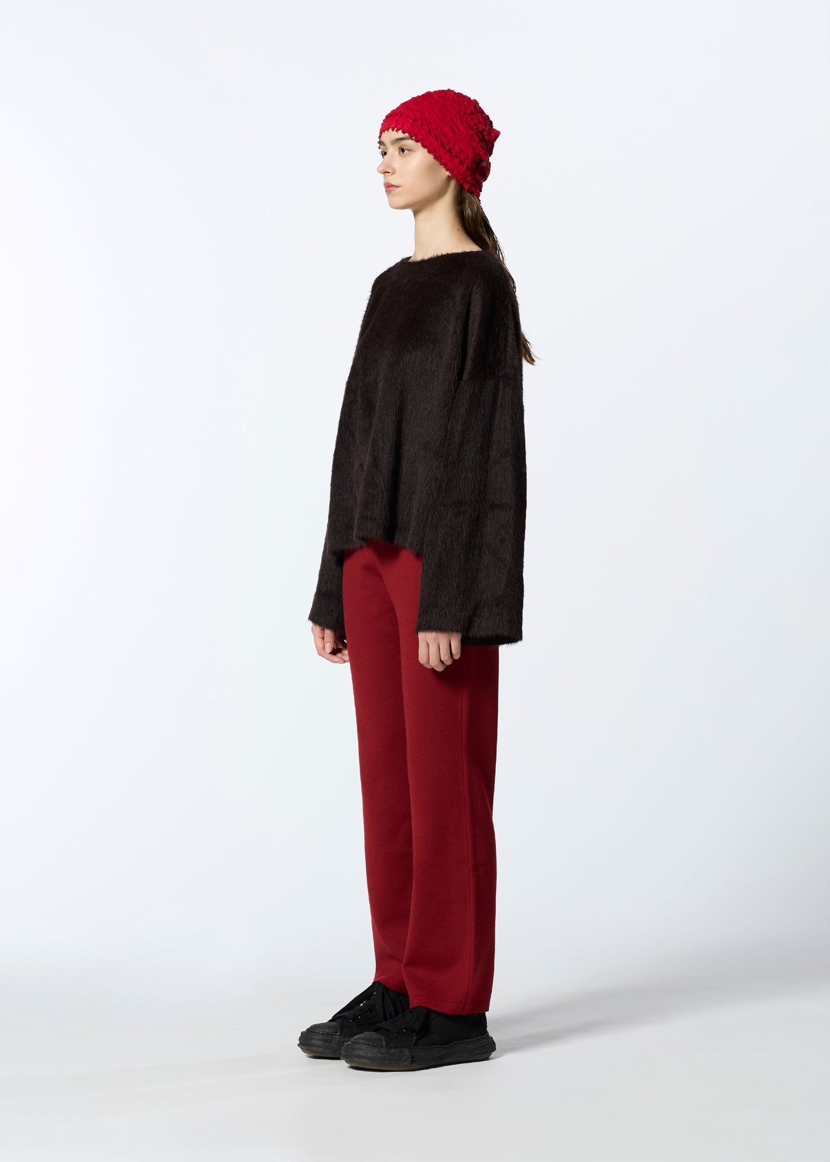 A person stands in profile against a white background, wearing HaaT’s SURI ALPACA dark brown wool sweater, wide red pants, black shoes, and a red knit beanie. view 6 of 7