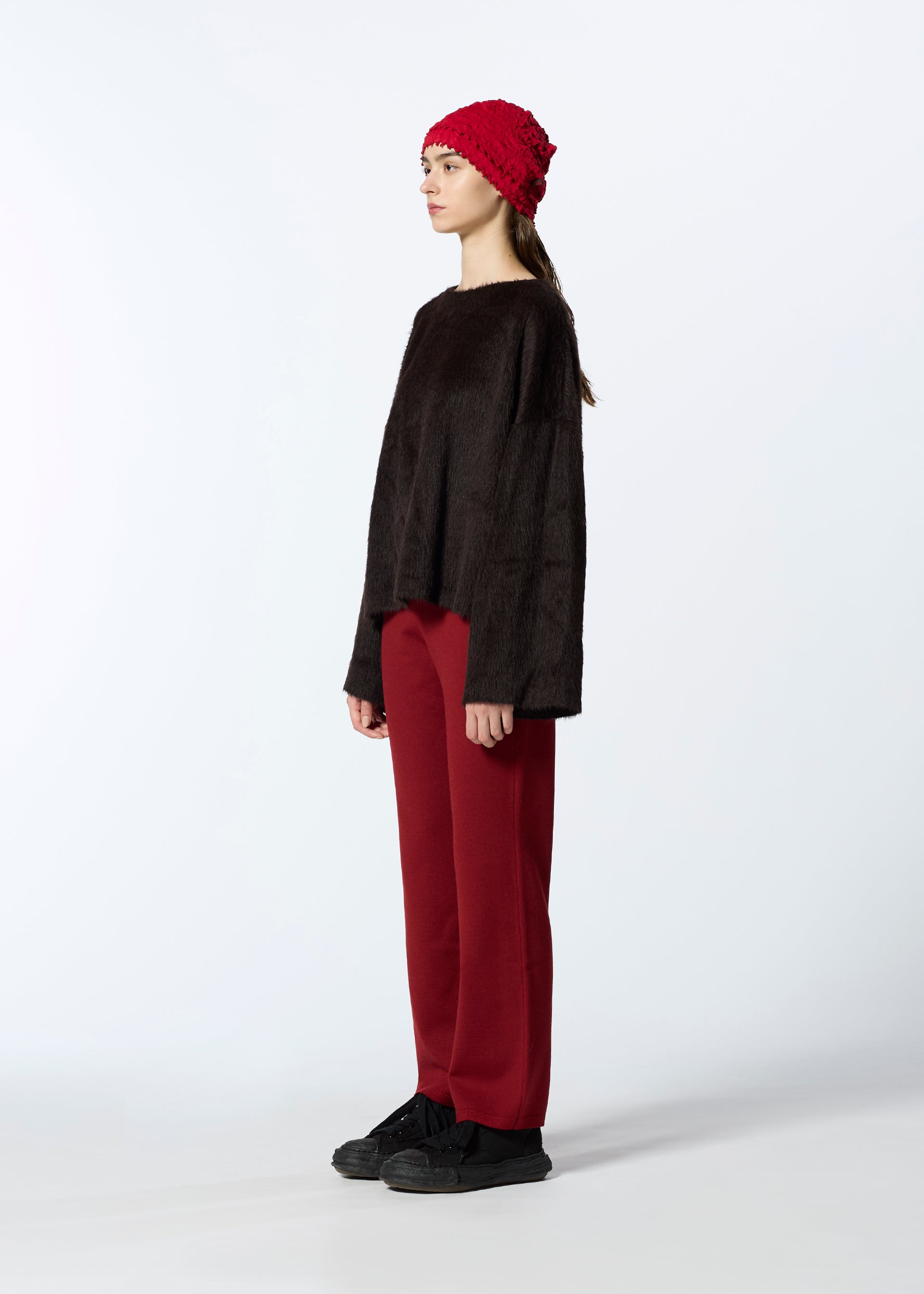 Zoomed view A person stands in profile against a white background, wearing HaaT’s SURI ALPACA dark brown wool sweater, wide red pants, black shoes, and a red knit beanie.