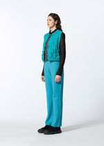 A person stands in profile against a plain background, wearing HaaT's 3D△ KNIT turquoise pants and matching vest, paired with a black long-sleeve top and black shoes.