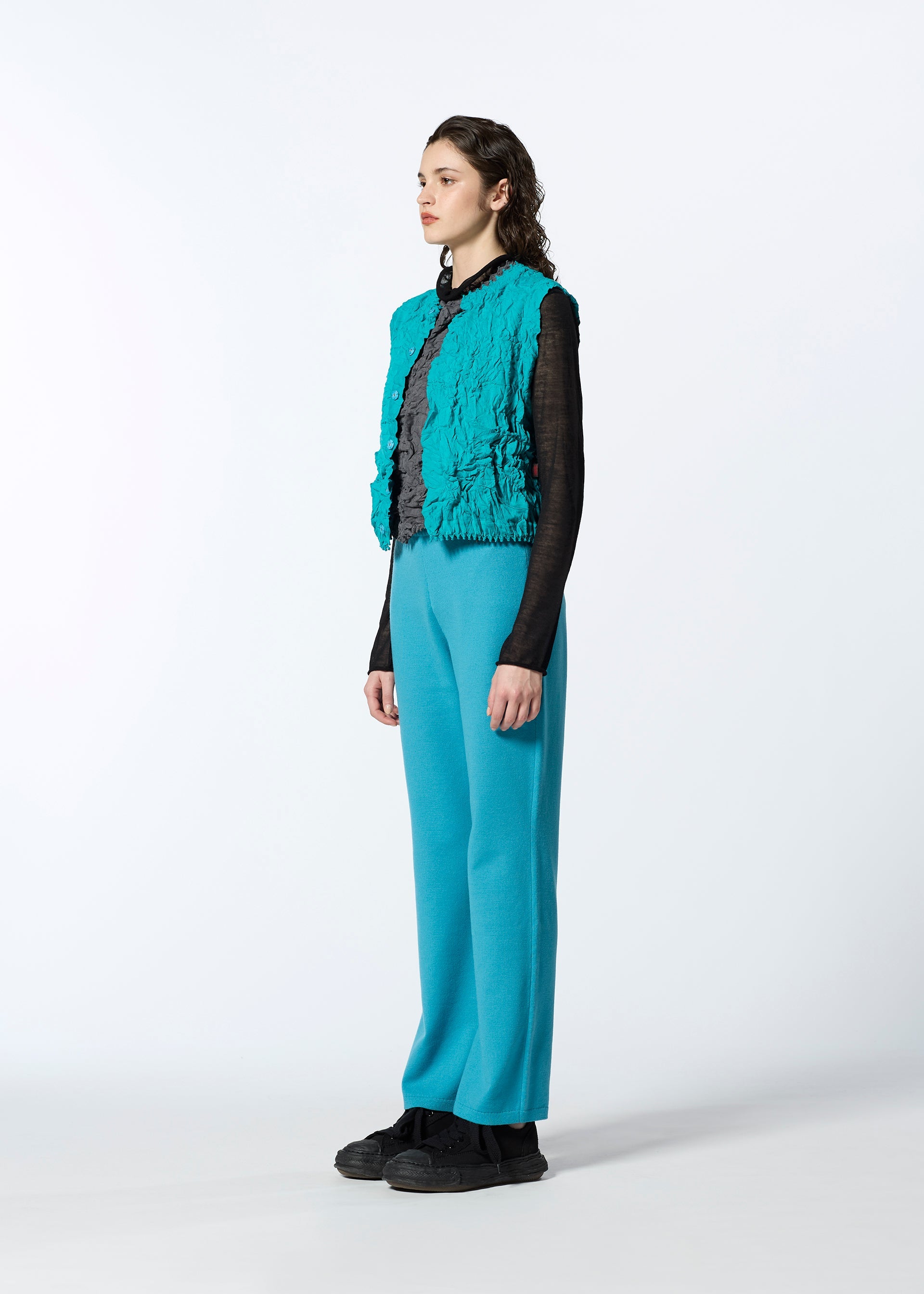 Zoomed view A person stands in profile against a plain background, wearing HaaT's 3D△ KNIT turquoise pants and matching vest, paired with a black long-sleeve top and black shoes.