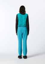 A person with curly hair faces away, wearing HaaT's 3D△ KNIT textured turquoise vest, wool-blend knit pants, a black long-sleeve top, and black shoes, standing against a plain light background.