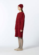 A woman stands sideways in HaaT's 3D△ KNIT—a loose, knee-length red knit dress with textured cutouts—paired with a matching beanie, light gray tights, and beige fuzzy slippers against a plain background.
