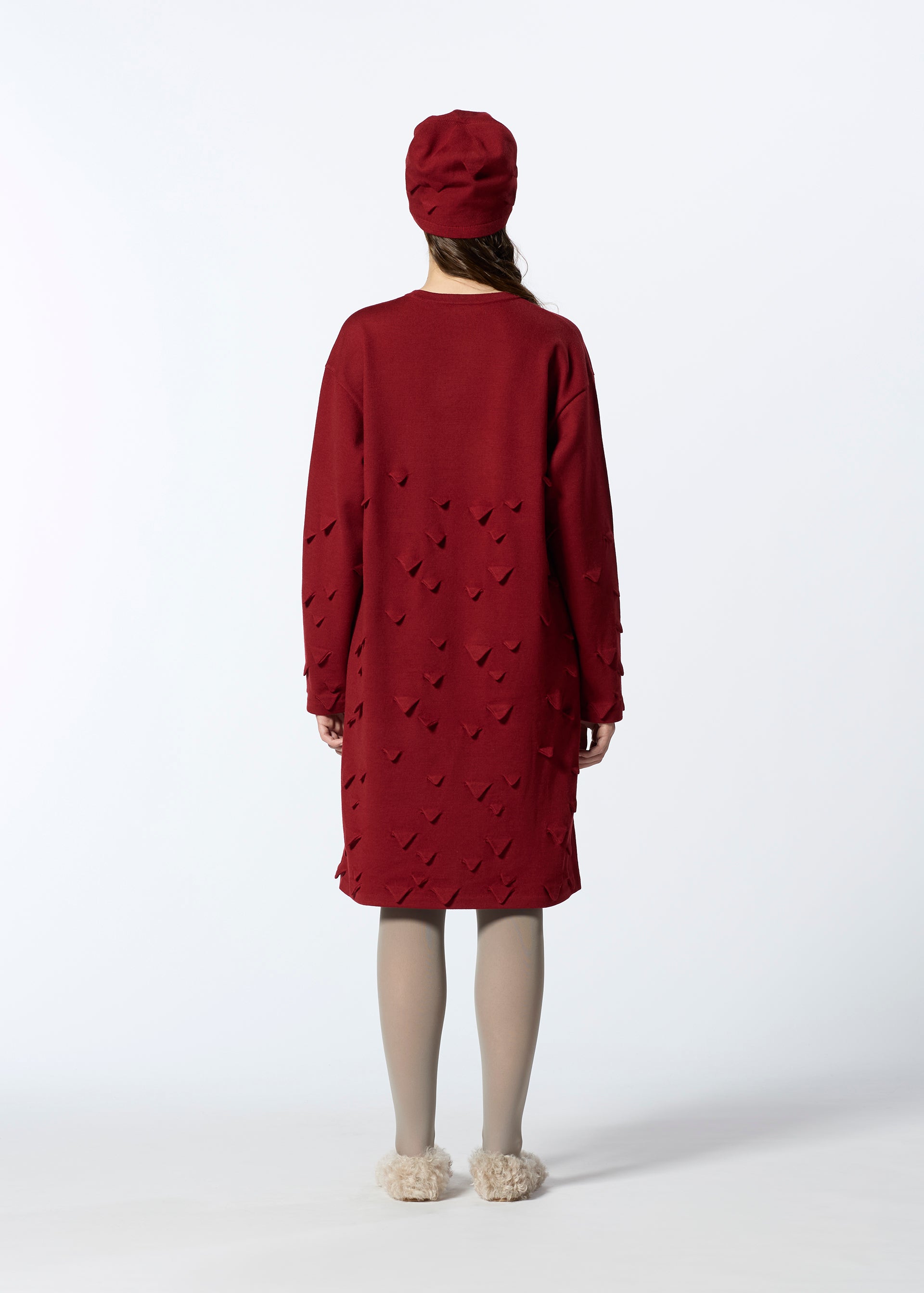 Zoomed view A person faces away, wearing HaaT's 3D△ KNIT long red wool-blend dress with cut-out details, a matching red hat, beige tights, and fluffy slippers against a plain white background.
