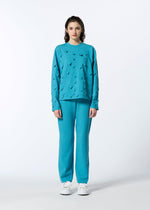 A person with shoulder-length dark hair stands against a white background, wearing the HaaT 3D△ KNIT wool-blend turquoise sweater with triangular cutouts, matching pants, and white sneakers.