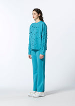 A person with wavy dark hair faces slightly left, wearing HaaT’s 3D△ KNIT turquoise long-sleeved cutout top and matching pants against a plain white background, paired with white sneakers.