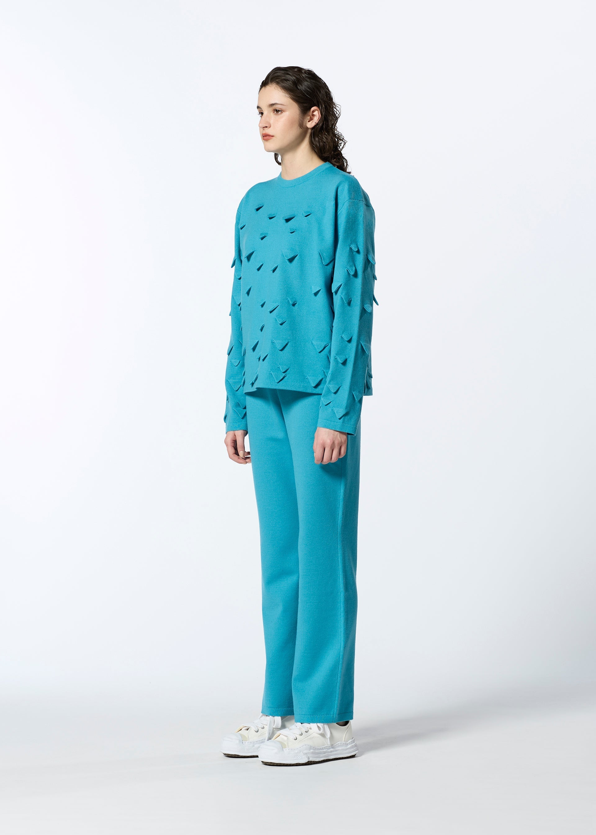 Zoomed view A person with wavy dark hair faces slightly left, wearing HaaT’s 3D△ KNIT turquoise long-sleeved cutout top and matching pants against a plain white background, paired with white sneakers.