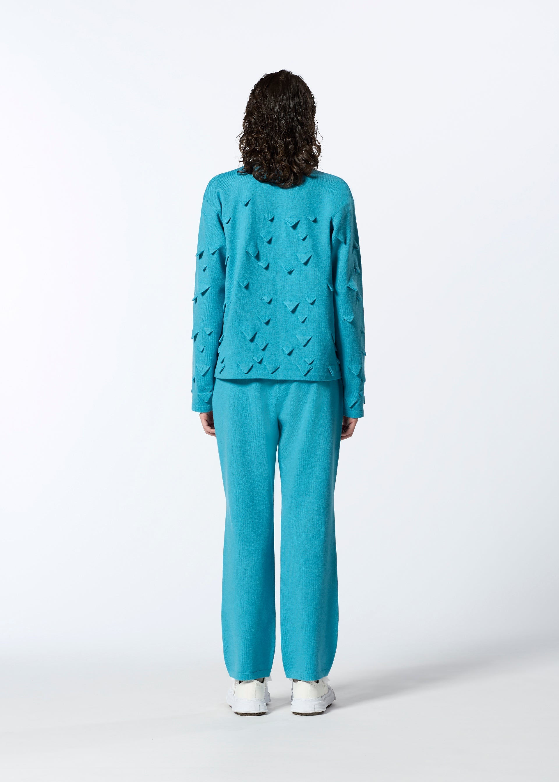 Zoomed view A person with curly hair faces away, wearing HaaT's 3D△ KNIT bright blue textured long-sleeved top and matching pants with white sneakers, set against a plain white background.