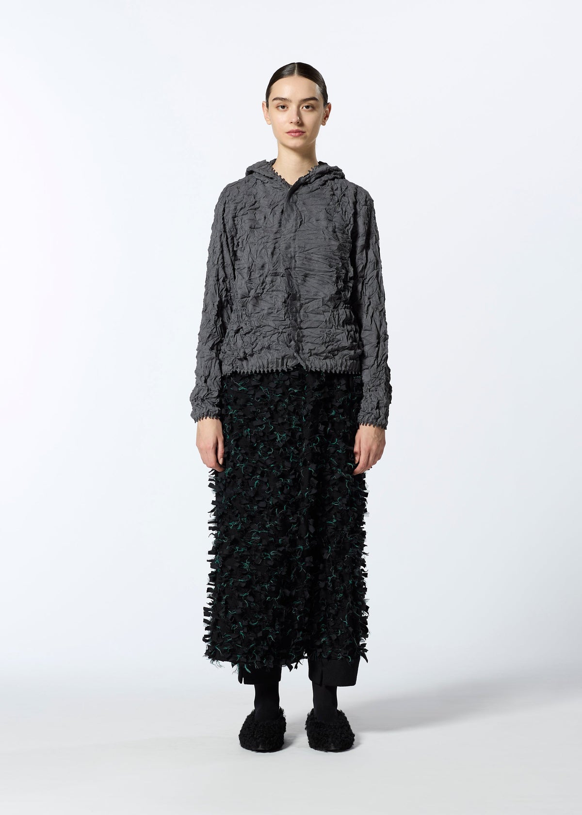 A person stands facing forward in a textured gray HaaT KUMO SHIBORI KNIT jacket, a long black patterned skirt, black pants, and black shoes against a plain white background. view 7 of 9