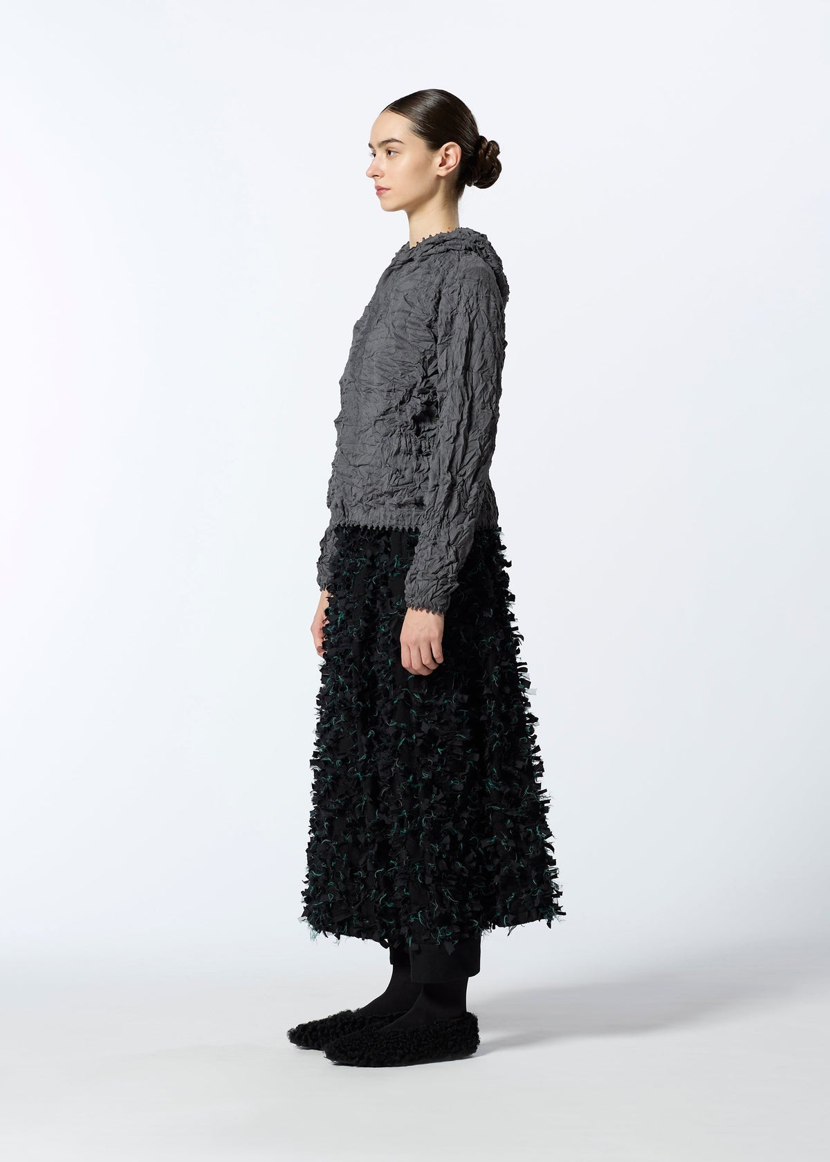 A woman in profile wears HaaT’s KUMO SHIBORI KNIT—a textured gray long-sleeve top made from recycled polyester—styled with a black ankle-length floral skirt, black socks, and textured black shoes, set against a plain white background. view 8 of 9