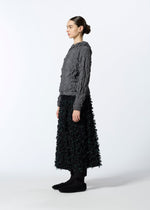 A woman in profile wears HaaT’s KUMO SHIBORI KNIT—a textured gray long-sleeve top made from recycled polyester—styled with a black ankle-length floral skirt, black socks, and textured black shoes, set against a plain white background.