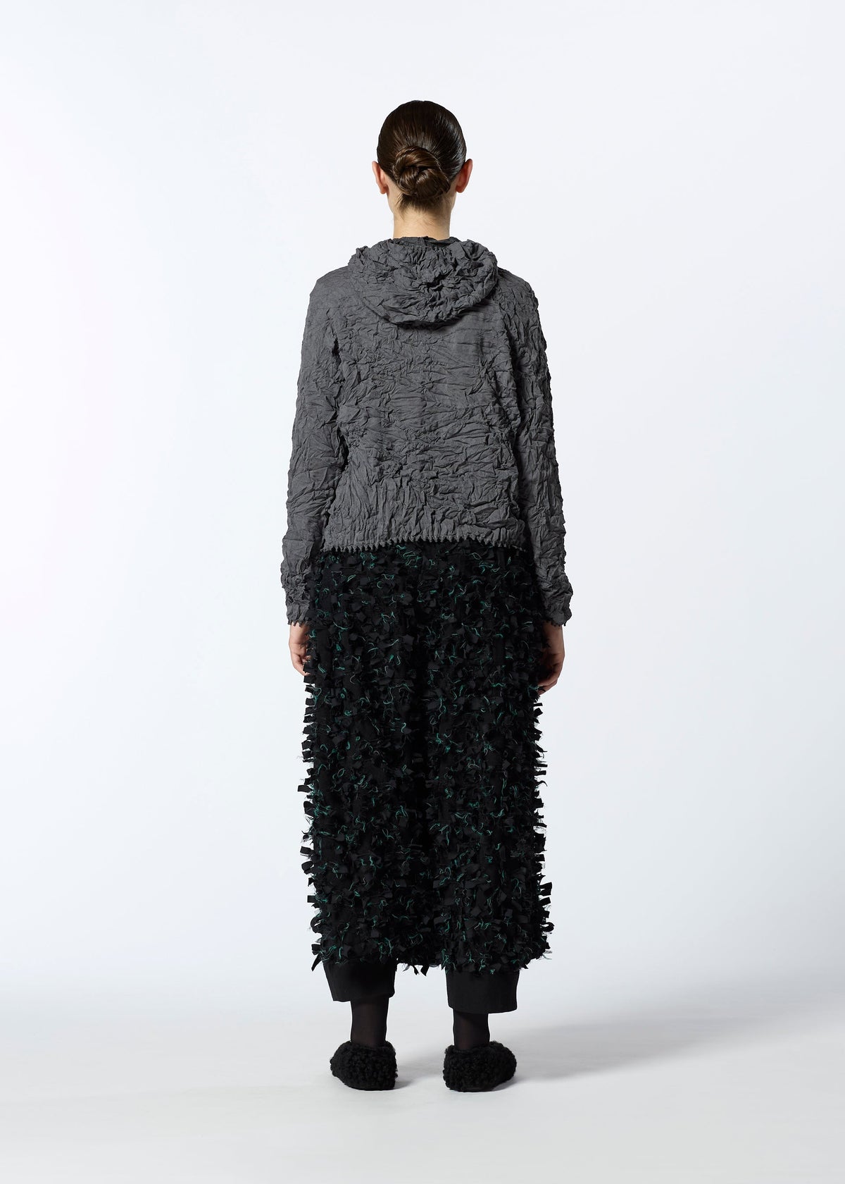 A person faces away wearing HaaT’s KUMO SHIBORI KNIT, a textured gray parka made from recycled polyester, with a black skirt featuring a fringed 3D pattern, paired with black leggings and shoes against a plain white background. view 9 of 9