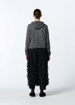 A person faces away wearing HaaT’s KUMO SHIBORI KNIT, a textured gray parka made from recycled polyester, with a black skirt featuring a fringed 3D pattern, paired with black leggings and shoes against a plain white background.