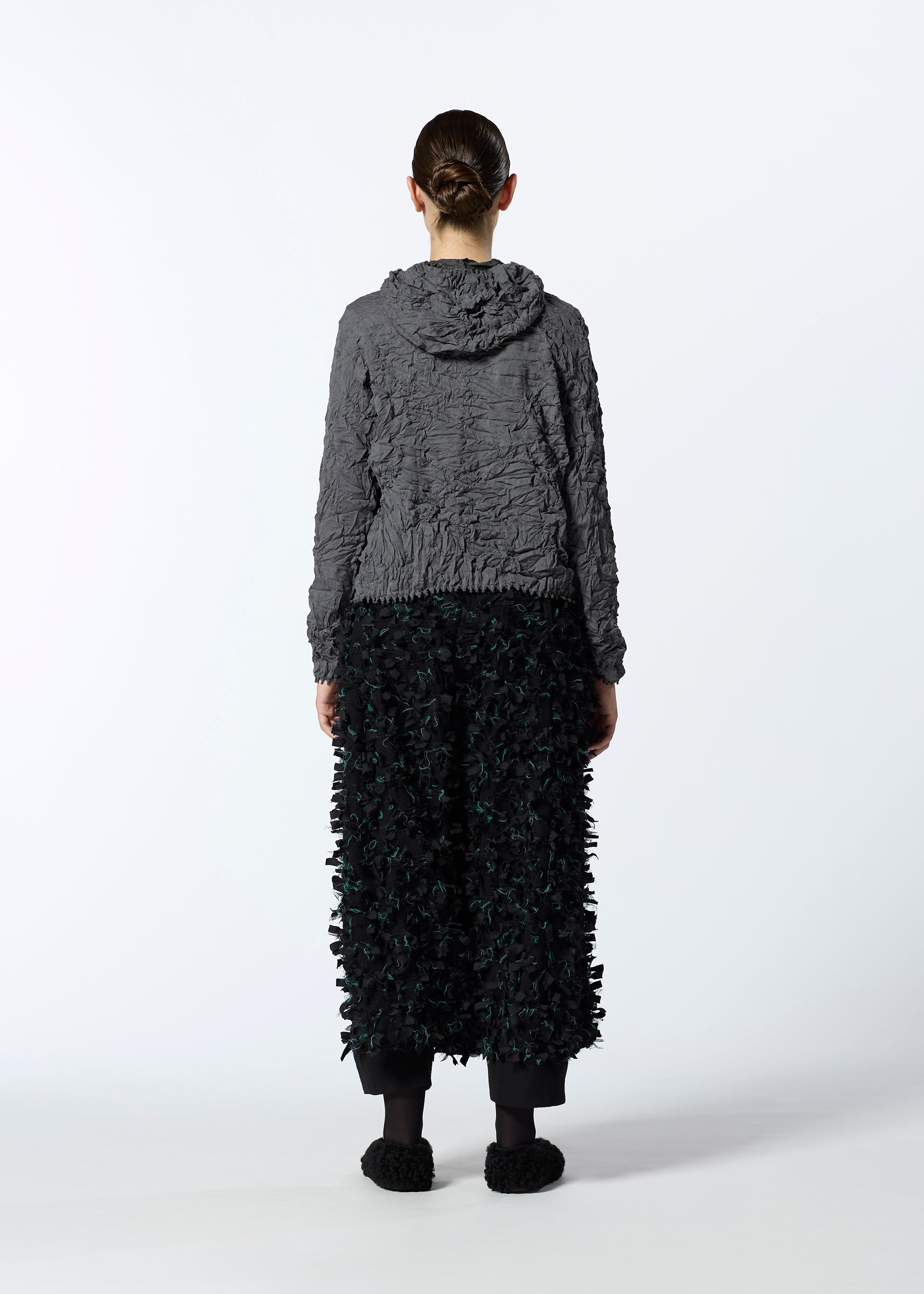 Zoomed view A person faces away wearing HaaT’s KUMO SHIBORI KNIT, a textured gray parka made from recycled polyester, with a black skirt featuring a fringed 3D pattern, paired with black leggings and shoes against a plain white background.