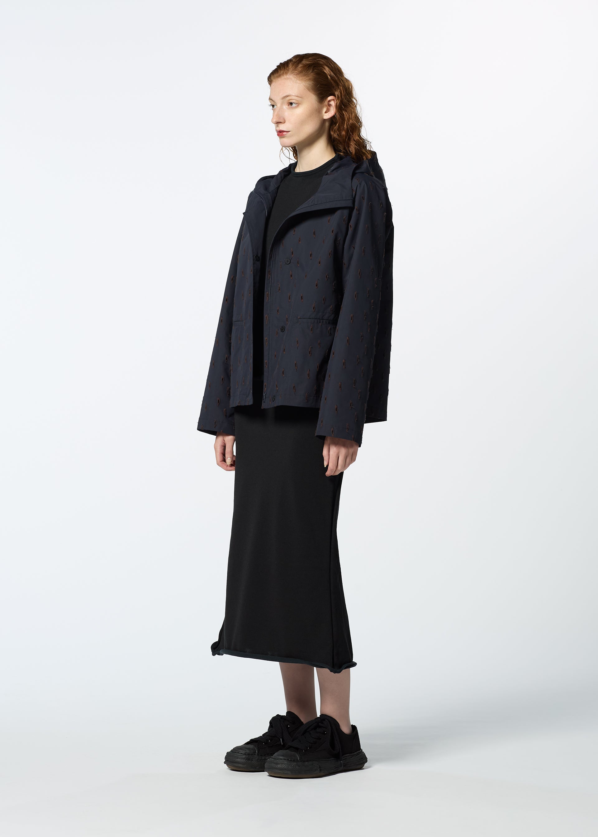 Zoomed view A light-skinned red-haired person in profile wears HaaT's TOBIKANNA dark hooded jacket with a subtle pattern, paired with a black ankle-length skirt and black sneakers, standing against a plain background.