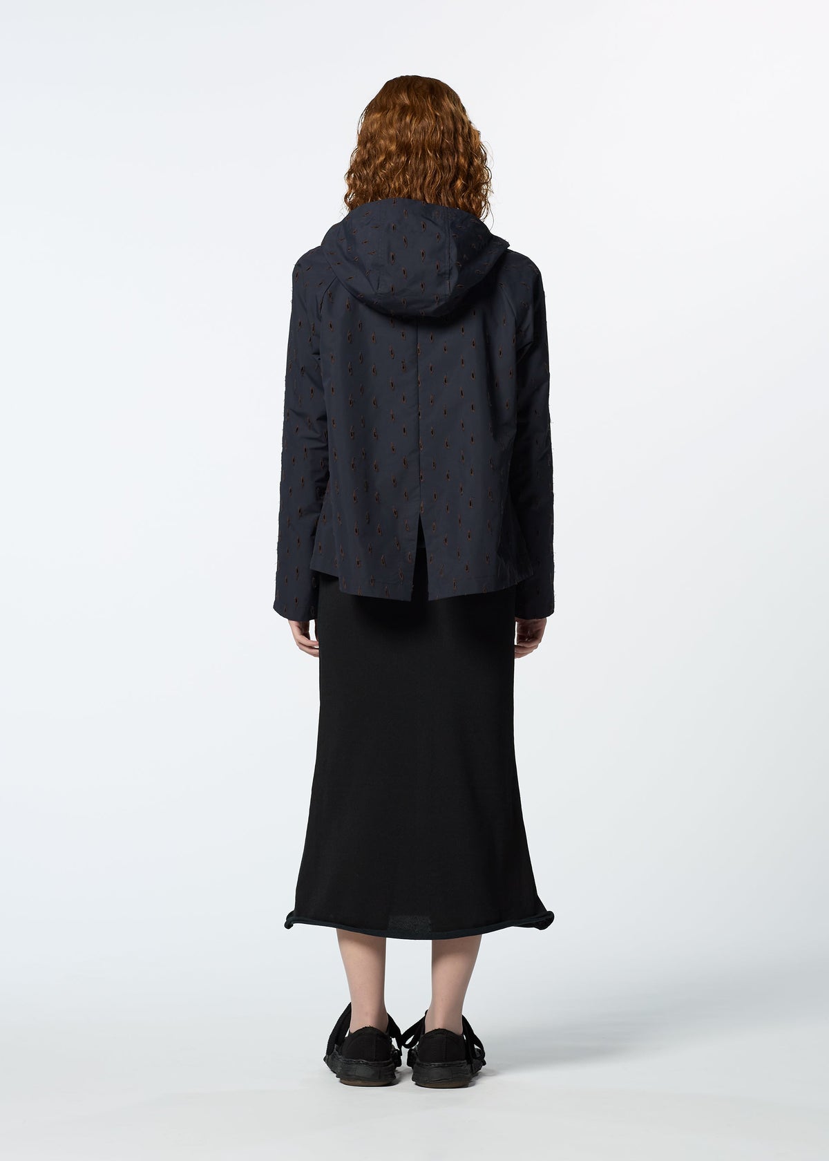 A person with curly hair, wearing HaaT's TOBIKANNA dark hooded jacket with subtle ceramic-inspired patterns and a long black skirt, stands facing away from the camera against a plain white background. view 9 of 11