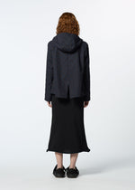 A person with curly hair, wearing HaaT's TOBIKANNA dark hooded jacket with subtle ceramic-inspired patterns and a long black skirt, stands facing away from the camera against a plain white background.