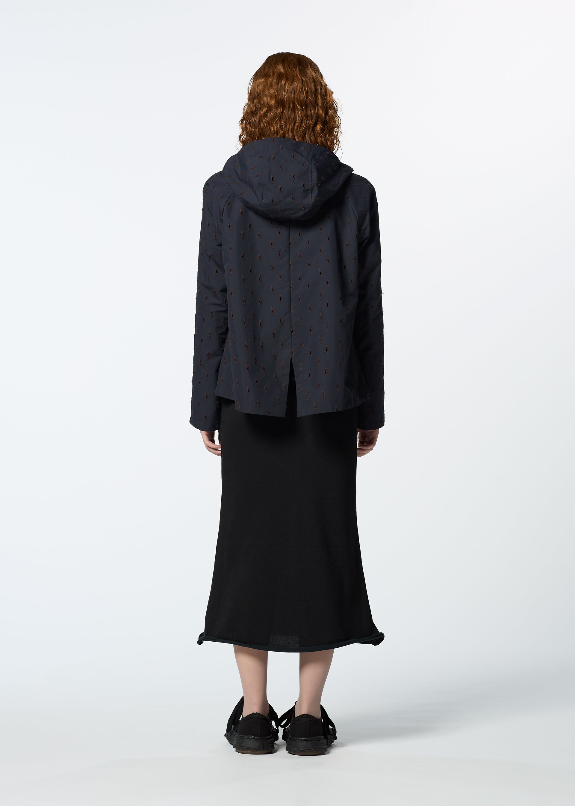 Zoomed view A person with curly hair, wearing HaaT's TOBIKANNA dark hooded jacket with subtle ceramic-inspired patterns and a long black skirt, stands facing away from the camera against a plain white background.