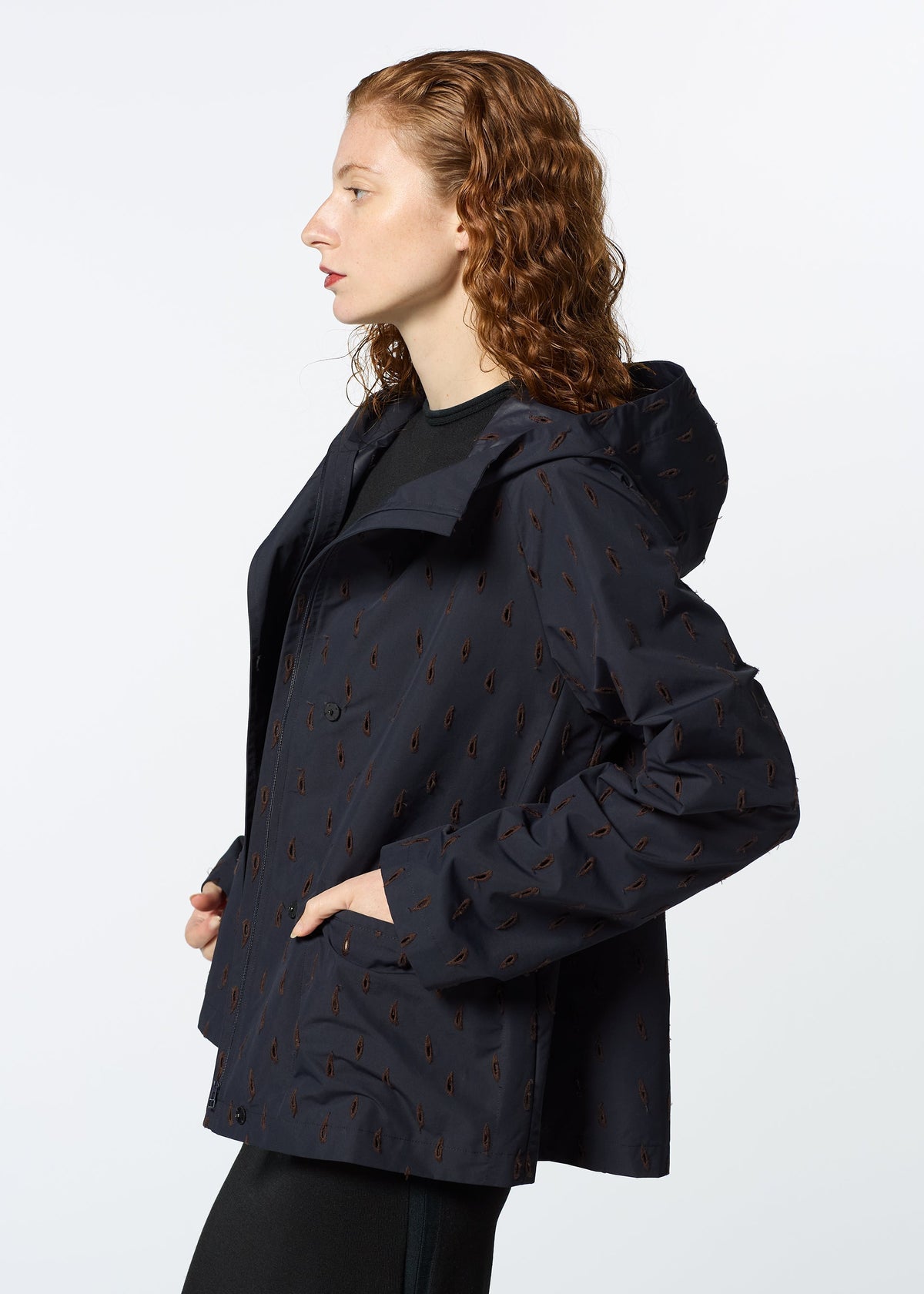 A woman with curly red hair wears HaaT’s TOBIKANNA dark hooded jacket, featuring a subtle pattern inspired by Tobikanna ceramics. She stands in profile, hands in pockets, against a plain white background. view 10 of 11