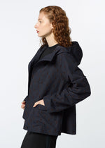 A woman with curly red hair wears HaaT’s TOBIKANNA dark hooded jacket, featuring a subtle pattern inspired by Tobikanna ceramics. She stands in profile, hands in pockets, against a plain white background.