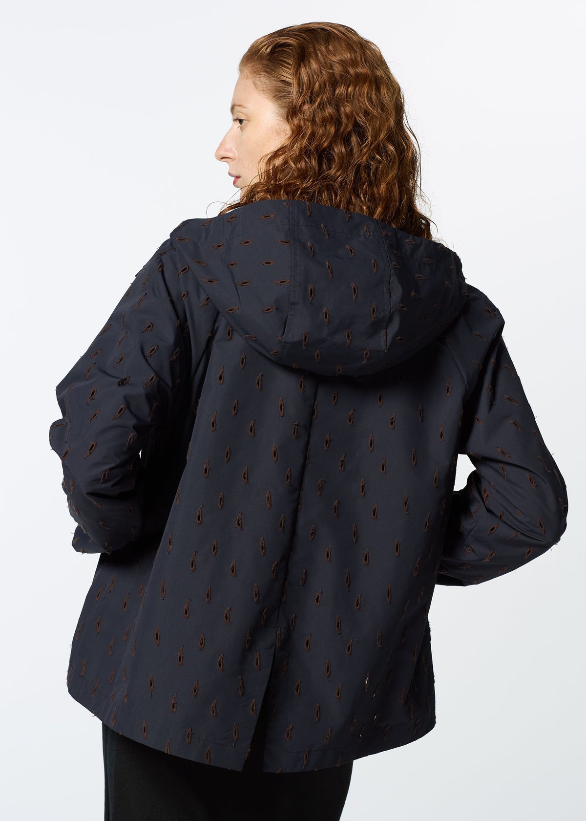 A person with wavy, reddish-brown hair is seen from behind, wearing HaaT’s TOBIKANNA dark navy hooded jacket featuring a subtle brown pattern inspired by ceramic designs, set against a plain light background. view 11 of 11
