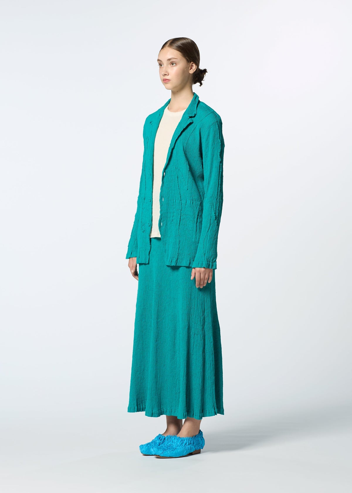 A person stands against a plain background, wearing a tailored teal KYO CHIJIMI EMB jacket and long skirt by HaaT with teal shoes and a cream top peeking underneath. Their hair is tied back as they face slightly to the side. view 8 of 9