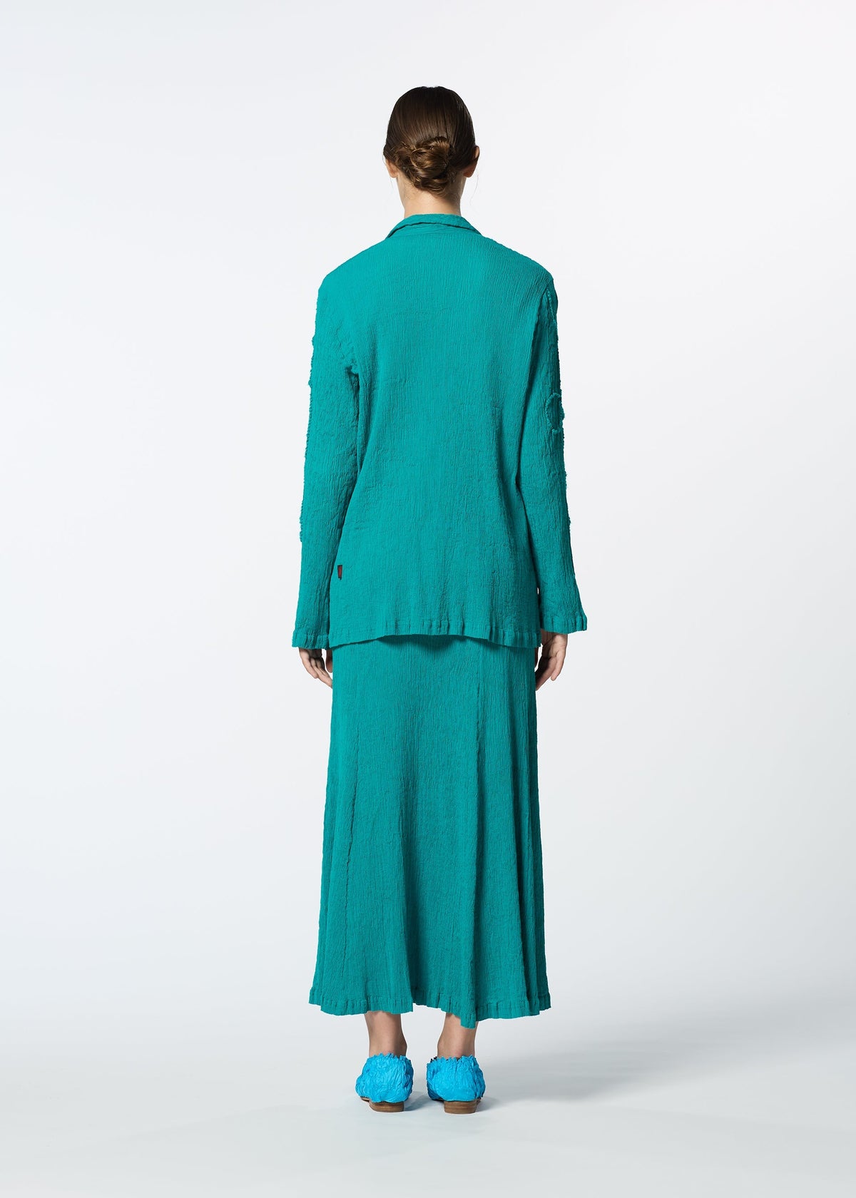 A person stands facing away in a KYO CHIJIMI EMB teal textured long blazer and matching ankle-length skirt by HaaT, paired with teal shoes, set against a plain white background. view 9 of 9