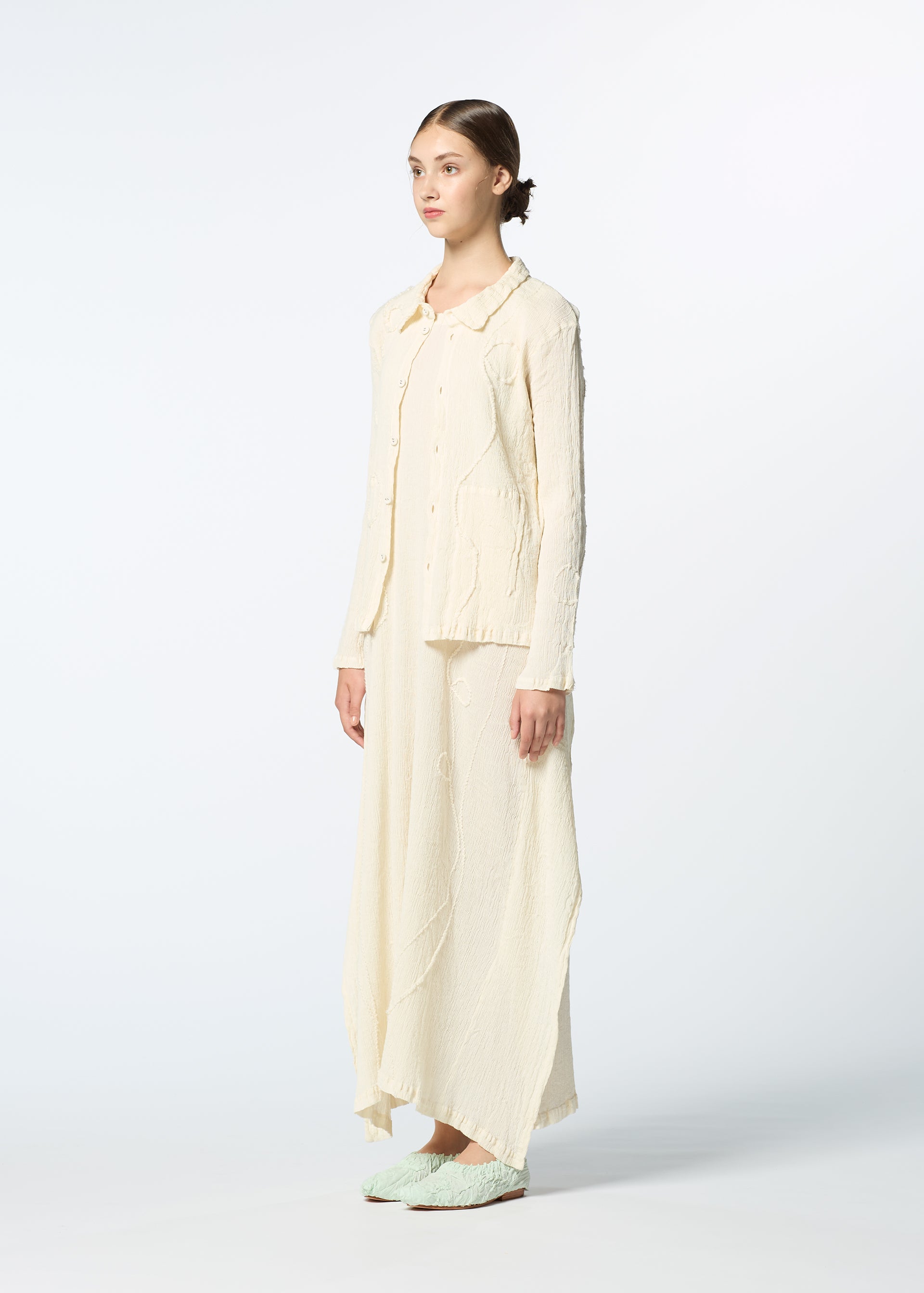 Zoomed view A woman stands in a studio, facing slightly to the side, wearing HaaT’s KYO CHIJIMI EMB—an off-white textured long-sleeve top with curved embroidery and a matching skirt—her hair pulled back against a plain background.